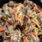 A fork lifts a serving of Keto Philly Cheese Steak Skillet—cooked beef strips, melted cheese, and sautéed red and green bell peppers—from a skillet filled with the same savory mixture.