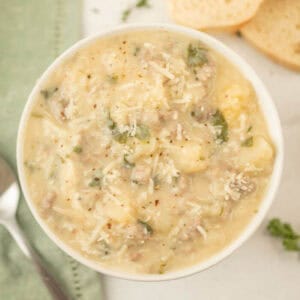 A bowl of creamy gnocchi soup reminiscent of a Copycat Olive Garden Zuppa Toscana Recipe, with visible parsley and cheese. It's accompanied by a spoon and slices of bread, appearing hearty with a light sprinkle of pepper on top.