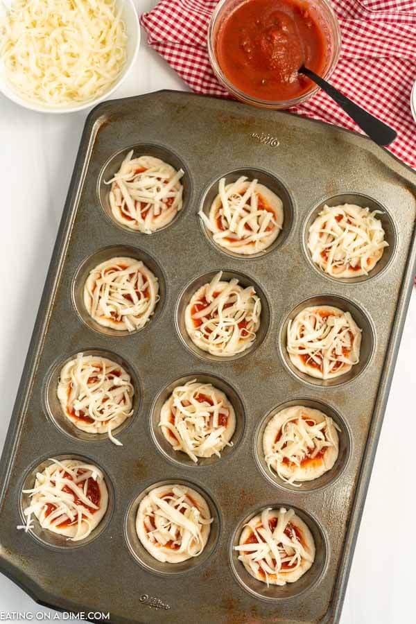 Uncooked pizza cupcakes in a muffin tin topped with cheese. 