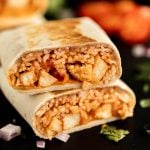 A close-up of a burrito cut in half, showing the delicious filling from a chicken burrito recipe—rice, chicken, beans, and sauce wrapped in a flour tortilla. The burrito is stacked on a dark surface with scattered onions and cilantro.