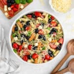 A large white bowl filled with Italian Pasta Salad—colorful rotini, cherry tomatoes, black olives, green bell peppers, cheese cubes, and shredded cheese—sits on a light surface, surrounded by wooden utensils and fresh ingredients.