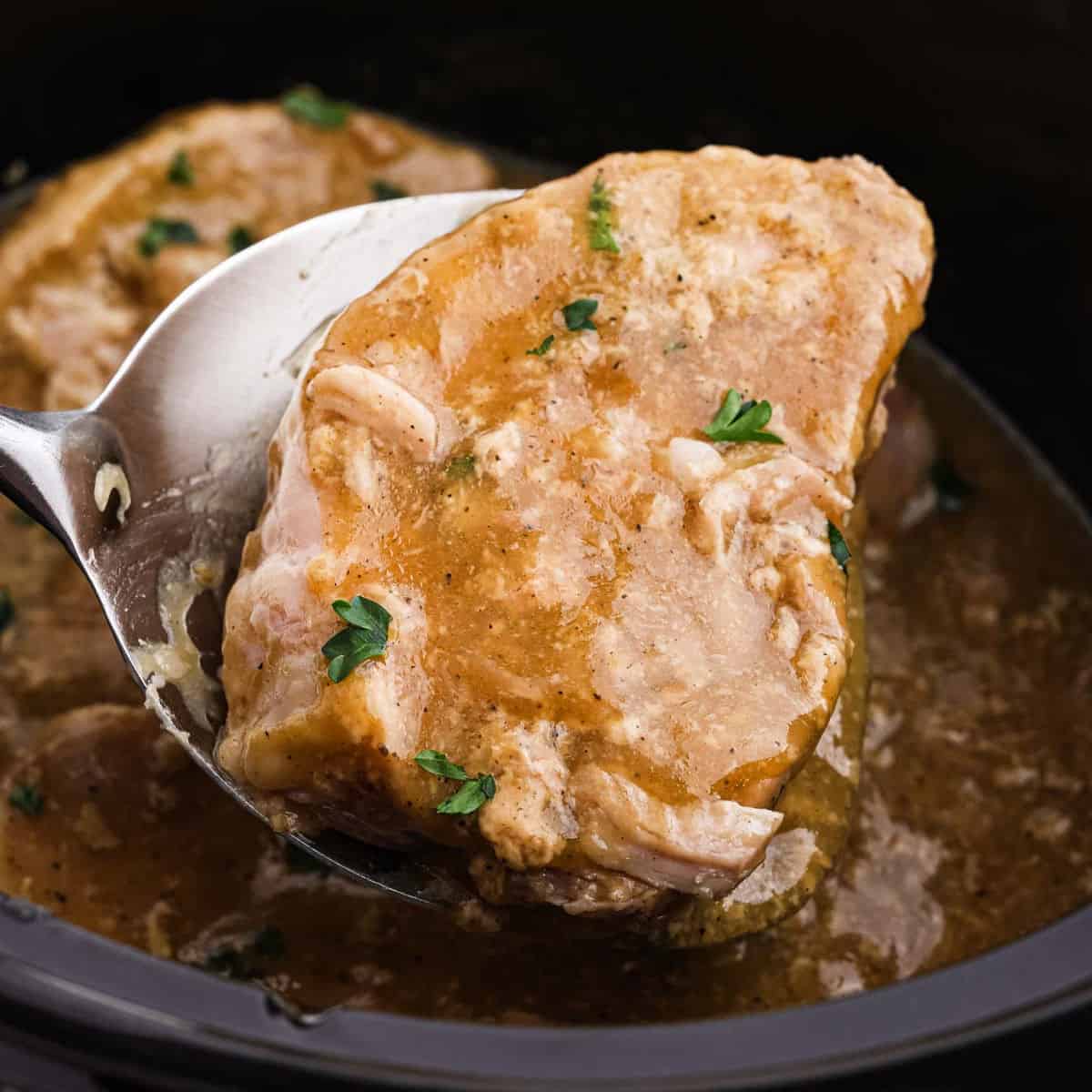 A close-up of a cooked pork chop being lifted from a slow cooker with a metal spatula, topped with sauce and garnished with fresh herbs—perfect for Crock Pot Pork Chops and Gravy lovers.