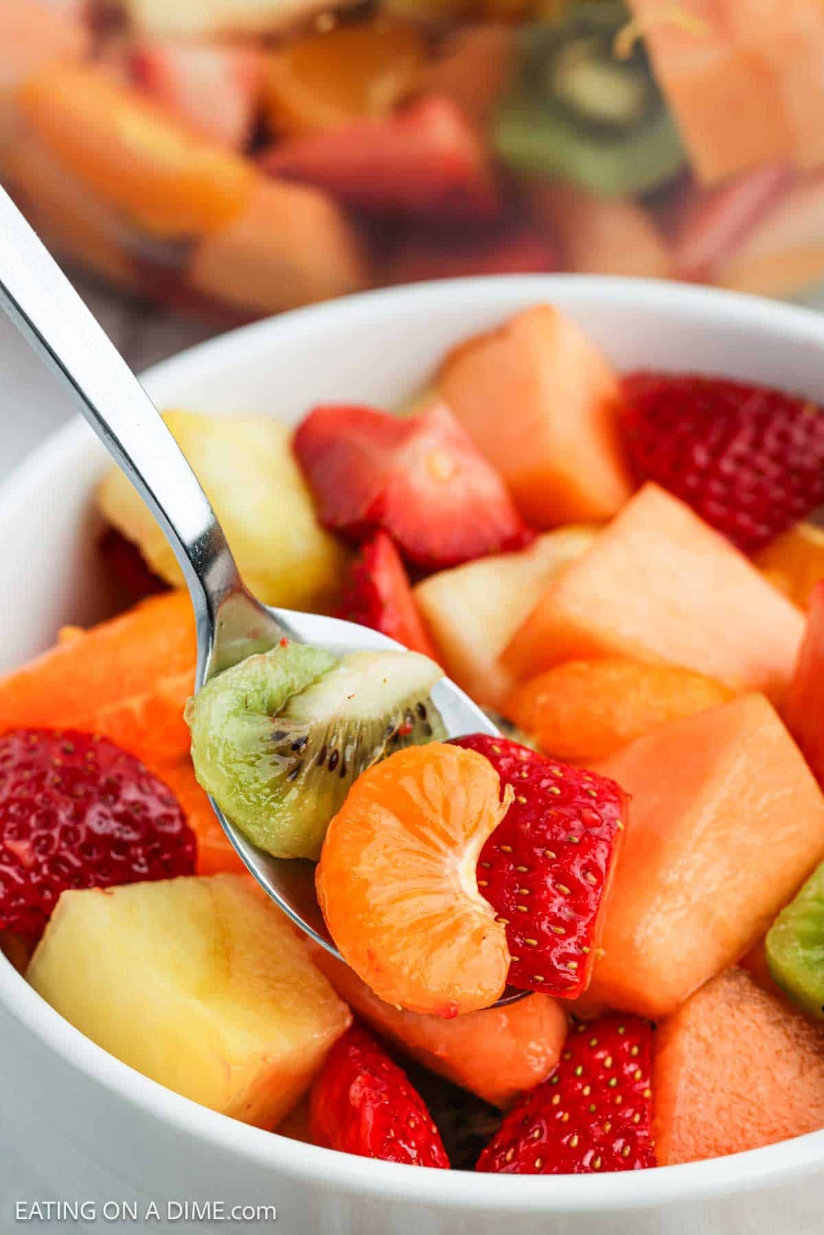 A mixture of fresh fruit in a bowl with a slice of kiwi, mandarin oranges and strawberries on a spoon.