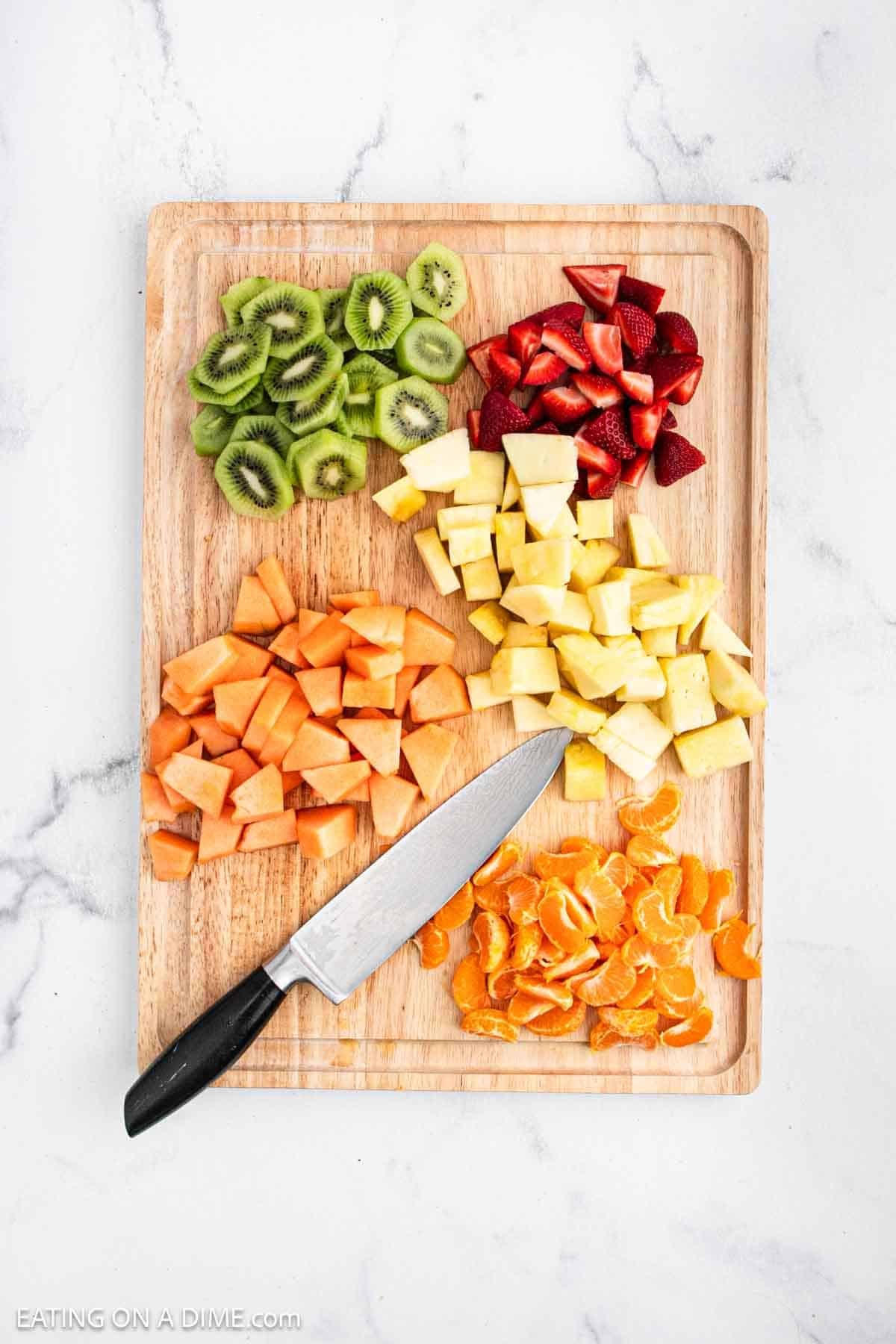 Fresh kiwi, strawberries, cantaloupe, oranges, and pineapple sliced on a cutting board with a knife.