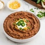 A bowl of Instant Pot homemade refried beans recipe topped with shredded cheese, sour cream, sliced jalapeños, and chopped green onions. Bowls of extra cheese and green onions are nearby on a white surface.