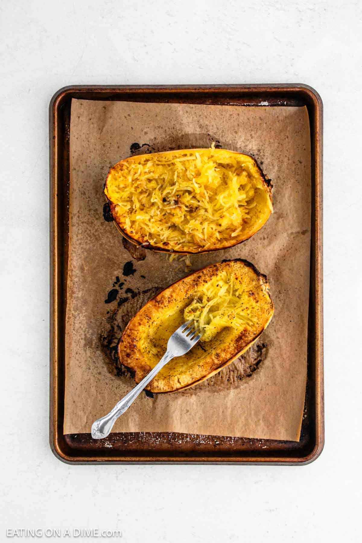 Cooked squash being shredding into strands with a fork on a baking sheet.