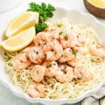A white plate of Shrimp Scampi featuring cooked shrimp on a bed of spaghetti, garnished with chopped parsley and served with two fresh lemon wedges.