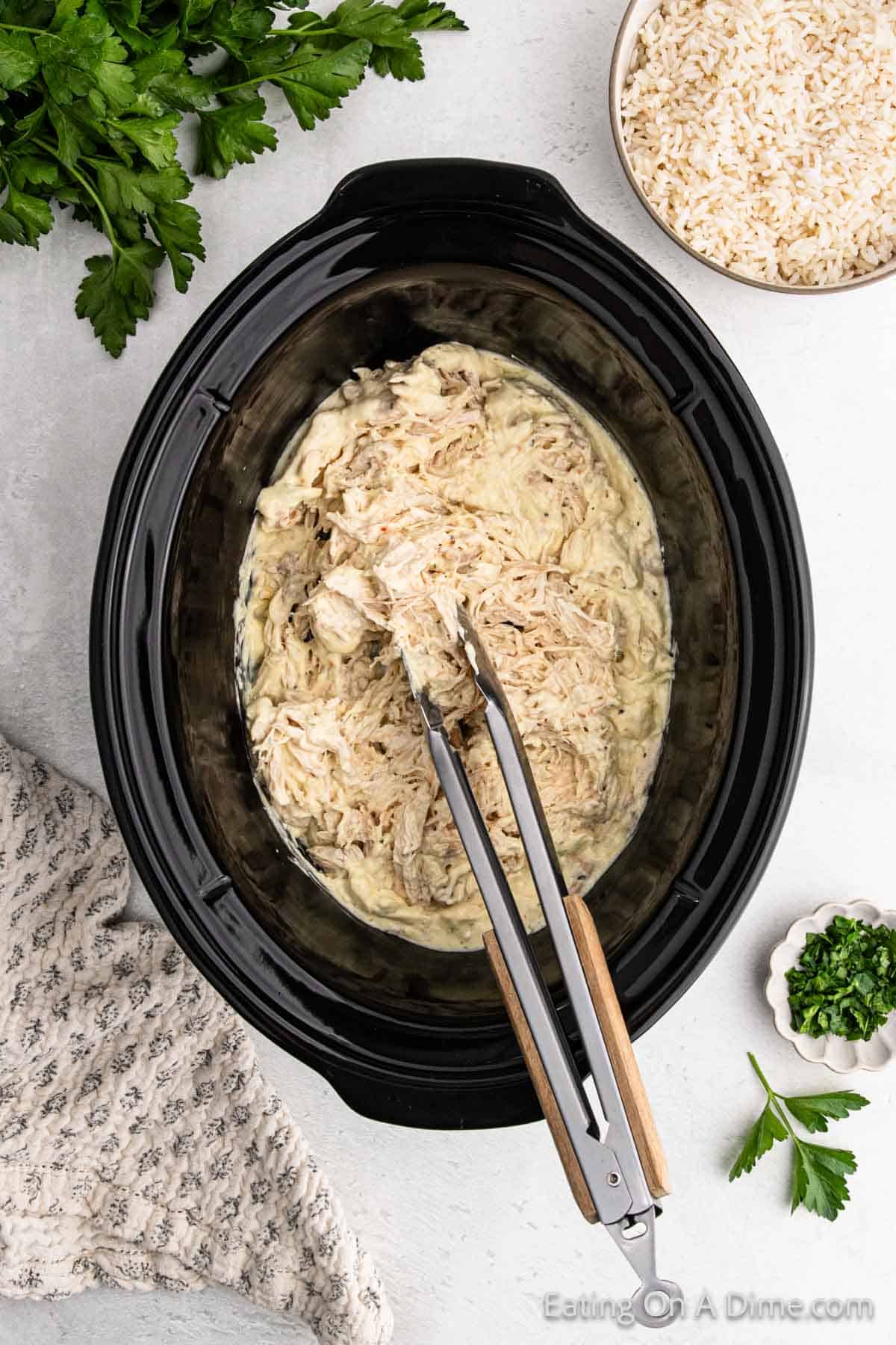 A slow cooker Italian chicken recipe featuring creamy shredded chicken awaits. Tongs rest on the savory mix, with fresh parsley and a bowl of rice nearby on the light countertop. A textured cloth adds a homely touch to the scene.