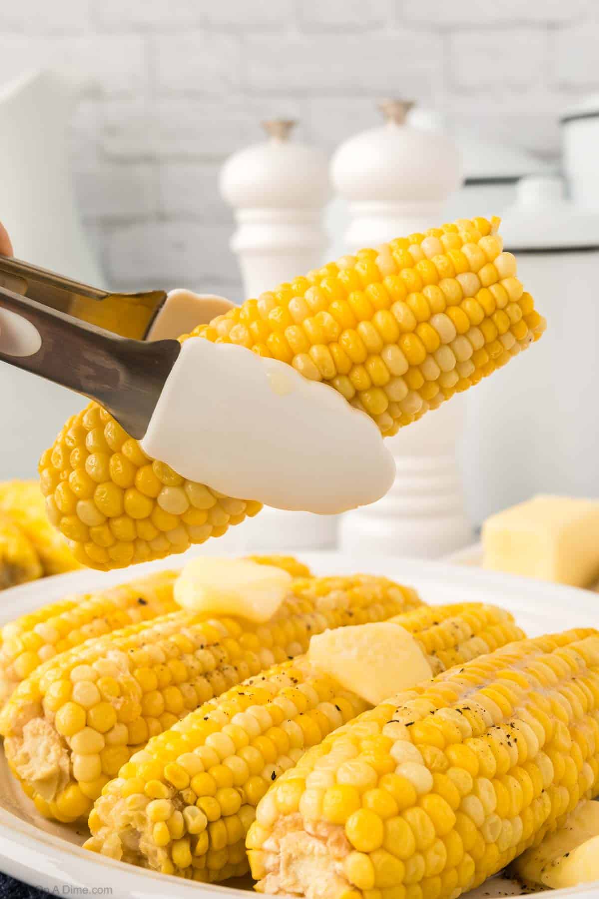 Corn on the cob between tongs with 4 corn on the cob on the platter topped with slice butter and salt and pepper
