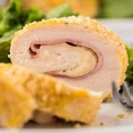 A close-up of a sliced Chicken Cordon Bleu recipe, showing layers of breaded chicken, ham, and melted cheese, with fresh green lettuce in the background.