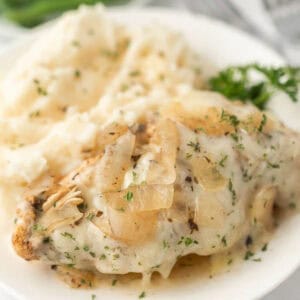 A plate with creamy mashed potatoes and Crockpot French Onion Chicken, topped with cooked onions and herbs, garnished with parsley.
