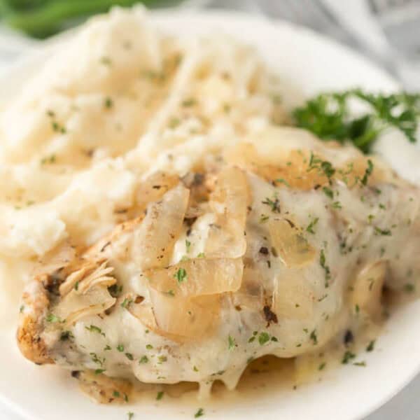A plate with creamy mashed potatoes and Crockpot French Onion Chicken, topped with cooked onions and herbs, garnished with parsley.