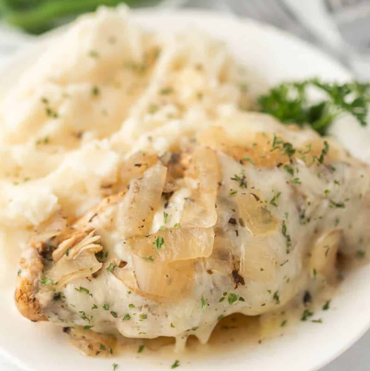 A plate with creamy mashed potatoes and Crockpot French Onion Chicken, topped with cooked onions and herbs, garnished with parsley.