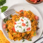 A white plate filled with cheesy Doritos Casserole nachos, topped with a dollop of sour cream and diced tomatoes, is sprinkled with chopped cilantro. A fork rests on the side, while bits of cilantro are visible in the background.