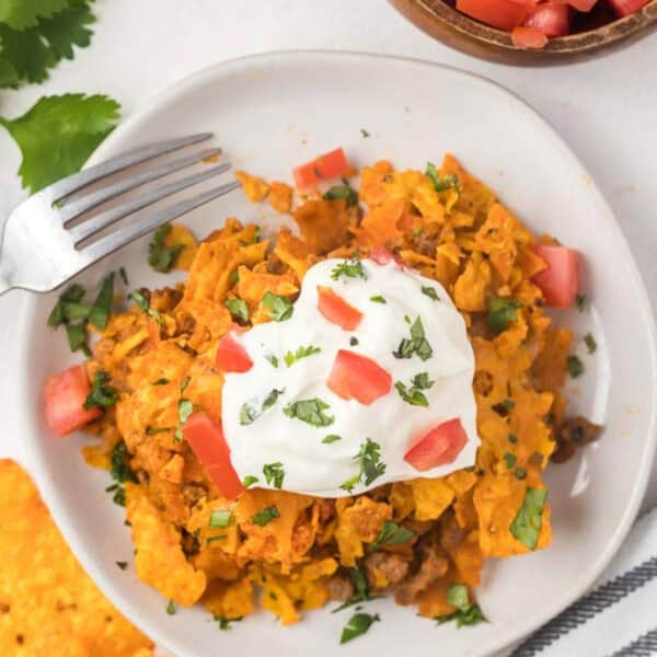 A white plate filled with cheesy Doritos Casserole nachos, topped with a dollop of sour cream and diced tomatoes, is sprinkled with chopped cilantro. A fork rests on the side, while bits of cilantro are visible in the background.