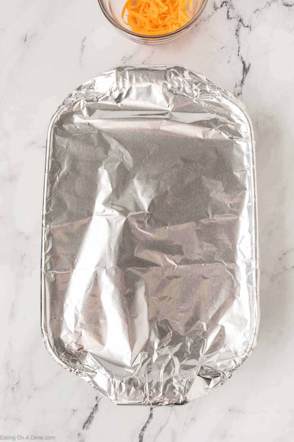 A rectangular baking dish covered with aluminum foil sits on a marble countertop. Above it is a small glass bowl of shredded cheddar cheese, perfect for topping your Easy Doritos Casserole Recipe.