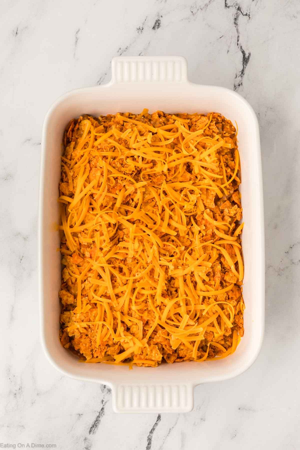 A white baking dish filled with a mixture of rice and ground meat, topped with shredded cheddar cheese, sits on a white marble surface—perfect for an Easy Doritos Casserole Recipe.