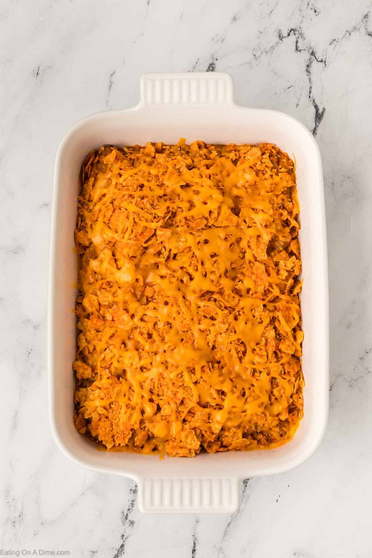 A rectangular white baking dish filled with an easy Doritos casserole recipe, topped with a layer of melted yellow cheese, sits on a white marble surface.