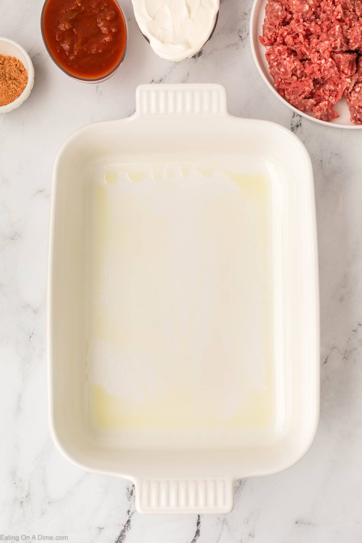 A greased white rectangular baking dish sits on a marble countertop, surrounded by bowls of ground meat, sour cream, salsa, and taco seasoning—perfect ingredients for an Easy Doritos Casserole Recipe.