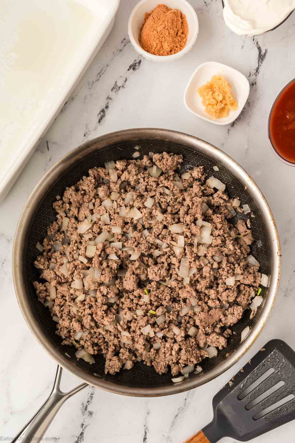 A frying pan on a marble counter holds cooked ground beef and diced onions—perfect for an Easy Doritos Casserole Recipe. Nearby are bowls with garlic, taco seasoning, and sour cream, plus a spatula and a dish of red sauce.