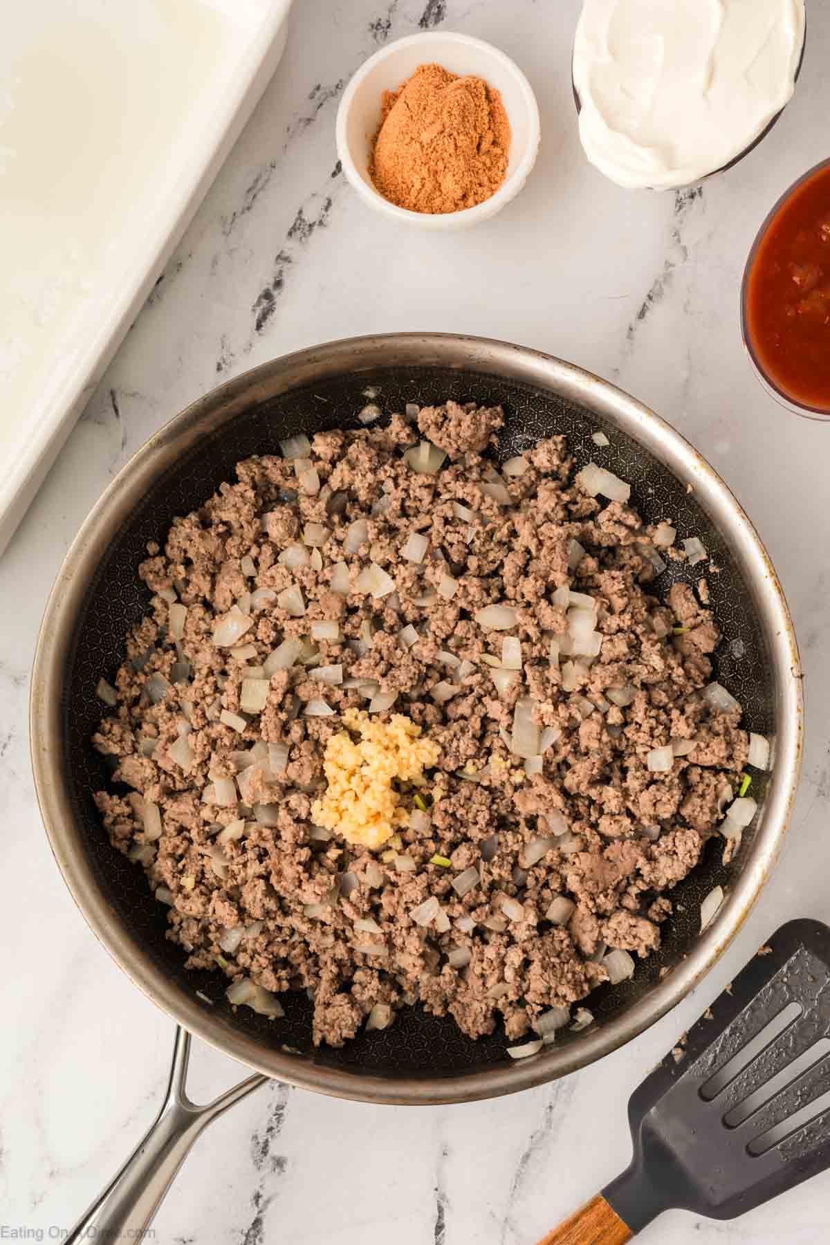 A skillet filled with cooked ground beef, chopped onions, and minced garlic sits on a marble counter—perfect for an Easy Doritos Casserole Recipe. Surrounding it are bowls of brown sugar, sour cream, and red sauce. A spatula lies nearby.