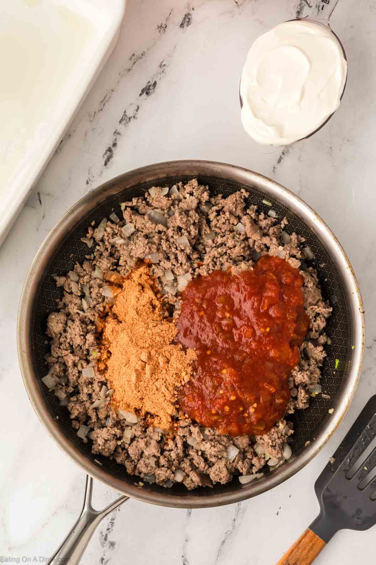 A skillet filled with cooked ground beef and diced onions, topped with taco seasoning powder and salsa, sits on a marble counter beside a bowl of sour cream and a spatula—perfect for an Easy Doritos Casserole Recipe.
