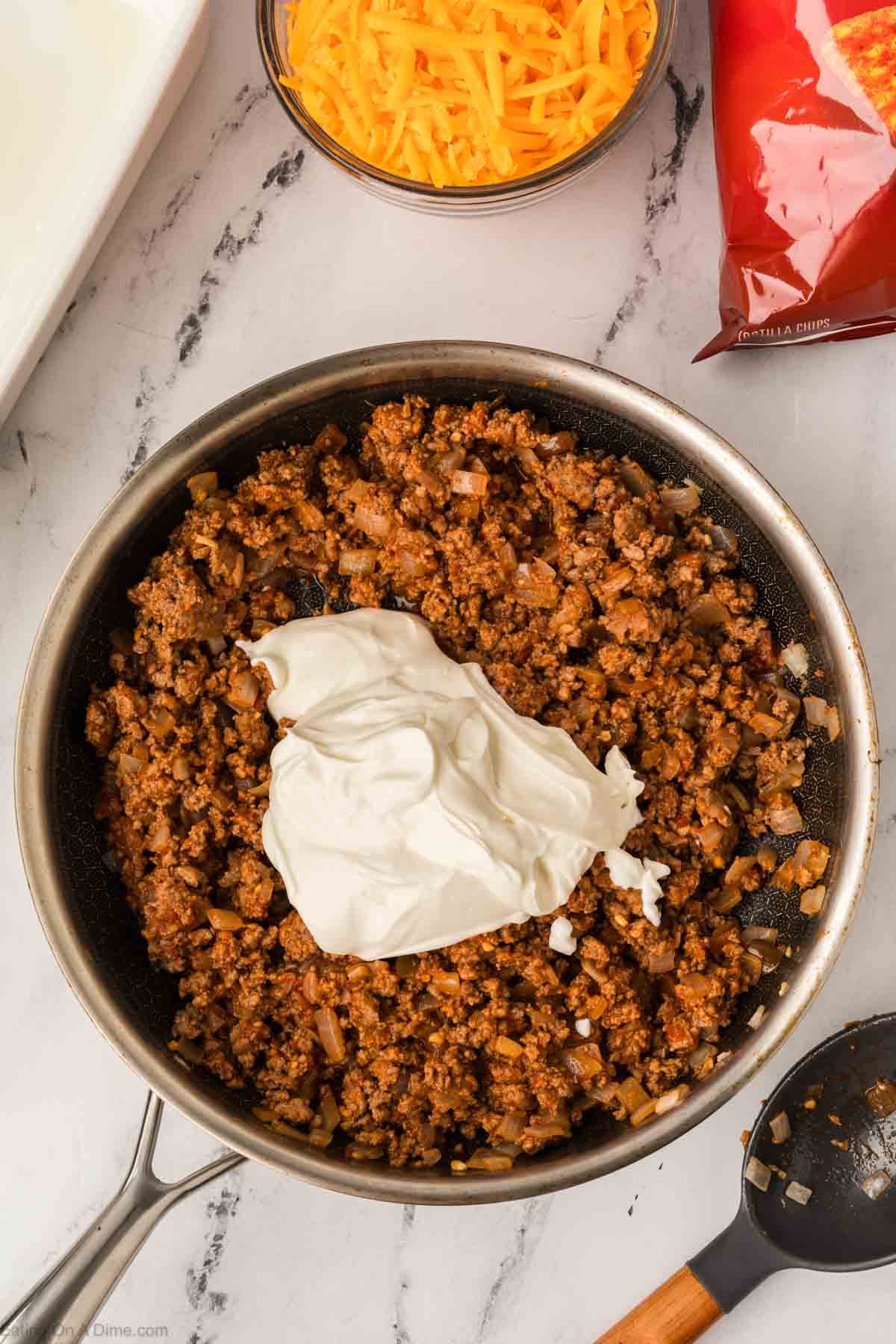 A skillet filled with cooked ground beef and onions, topped with a large dollop of sour cream. Nearby, a bowl of shredded cheddar cheese and a red tortilla chips bag hint at an Easy Doritos Casserole Recipe on a marble countertop.