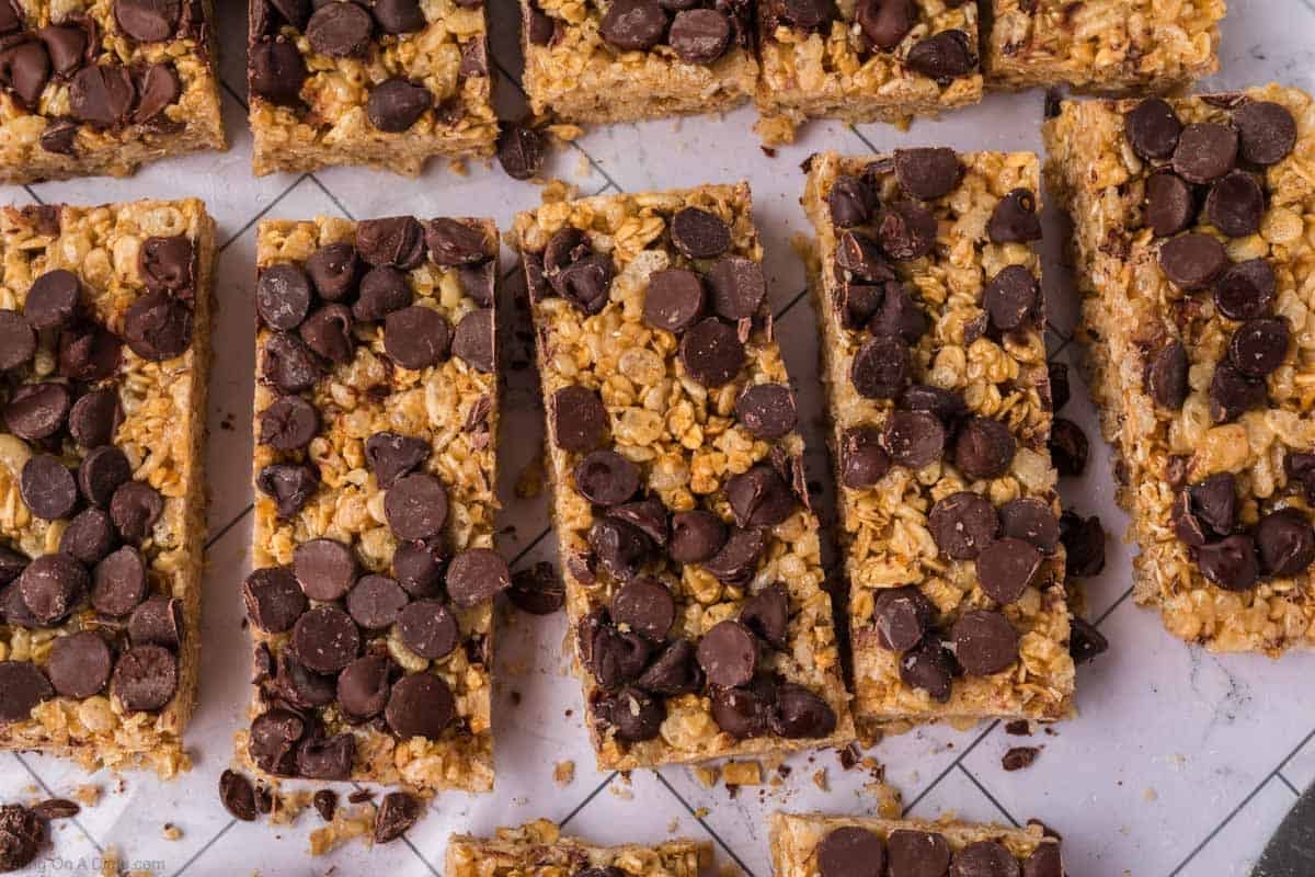 A close-up of homemade granola bars topped with chocolate chips, cut into rectangular pieces and arranged on a white surface. These delicious homemade granola bars feature oats and other visible grains for a wholesome snack.