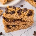 Three homemade granola bars stacked on top of each other, topped with chocolate chips, sit on a white surface with more homemade granola bars and crumbs scattered in the background.