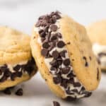 A close-up of a chocolate chip cookie ice cream sandwich with vanilla ice cream nestled between two soft and chewy cookies. The edges of the ice cream are coated in a layer of mini chocolate chips.