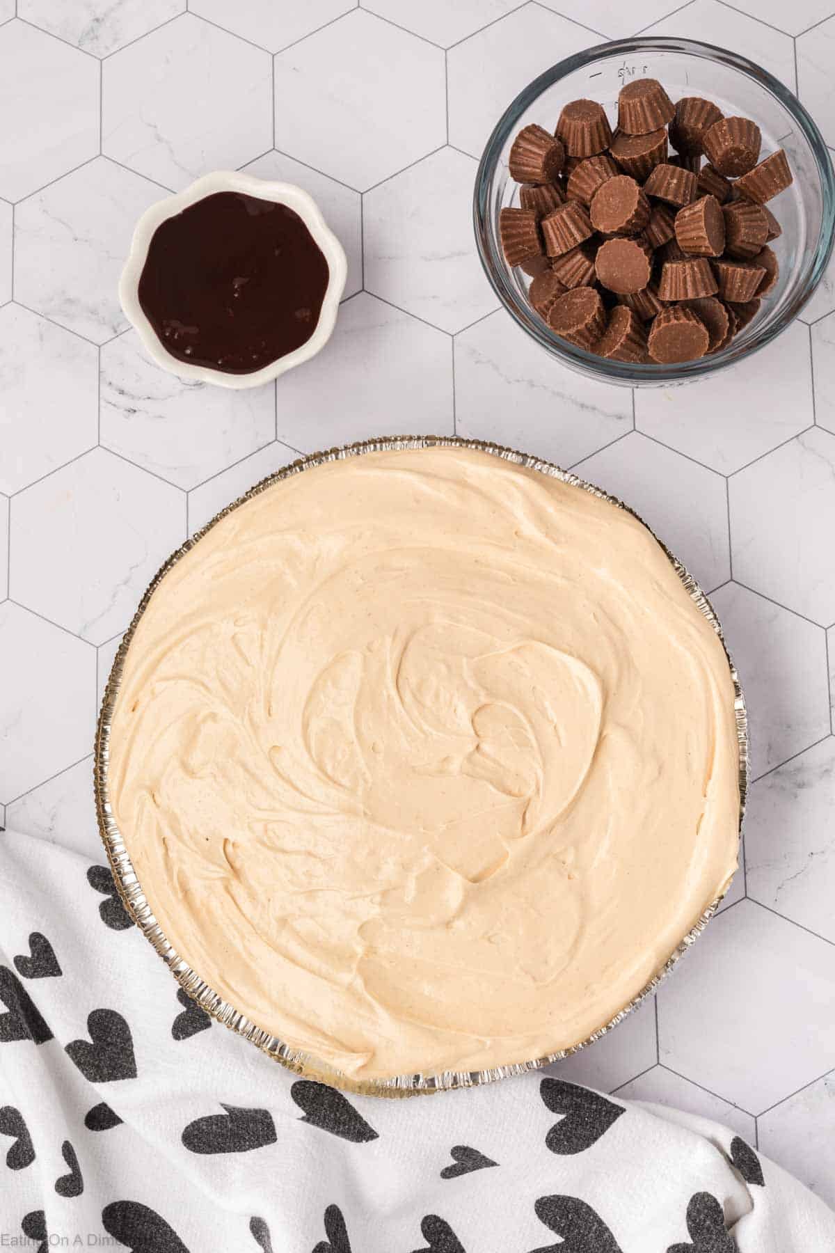 Peanut butter mixture placed in graham cracker crust with small bowl of chocolate syrup and mini peanut butter cups
