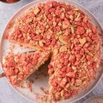 A round Strawberry Shortcake Cheesecake crumble cake with a slice being lifted out, topped with red and tan crumbly bits, displayed on a white plate.