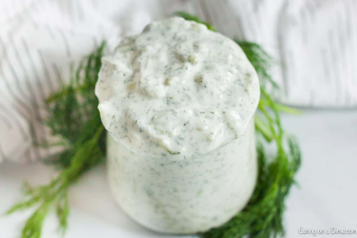 We have a really easy recipe for homemade tzatziki sauce that is amazing. Learn how to make tzatziki sauce and see just how easy and delicious it is! A jar filled with creamy tzatziki sauce, topped with chopped herbs, surrounded by fresh dill on a light surface. This image visually represents a tzatziki sauce recipe.
