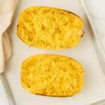 Two halves of cooked spaghetti squash, perfect for learning how to roast spaghetti squash, rest on a baking sheet with strands resembling spaghetti. Lightly seasoned with pepper, their vibrant texture is enticing. A white cloth is partially visible on the side, adding a touch of elegance.
