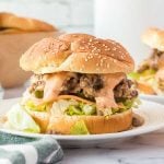 A sesame seed hamburger bun filled with Big Mac Sloppy Joes, shredded lettuce, cheese, pickles, and creamy sauce, served on a white plate with a blurred burger in the background.