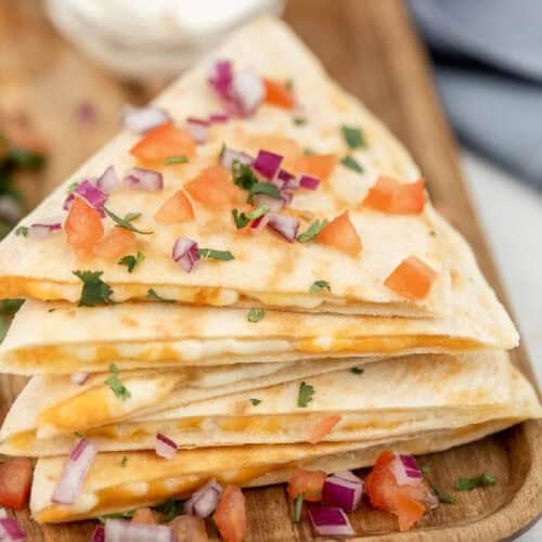 A stack of cheesy quesadillas, crafted from a delicious cheese quesadilla recipe, is cut into triangles and garnished with diced tomatoes, red onions, and cilantro on a wooden board. A small bowl of sour cream is visible in the background.