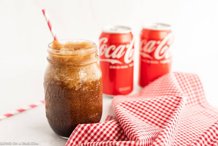 Close up image of a coke slushie in a mason jar with a straw and two cans of coca-cola in the background
