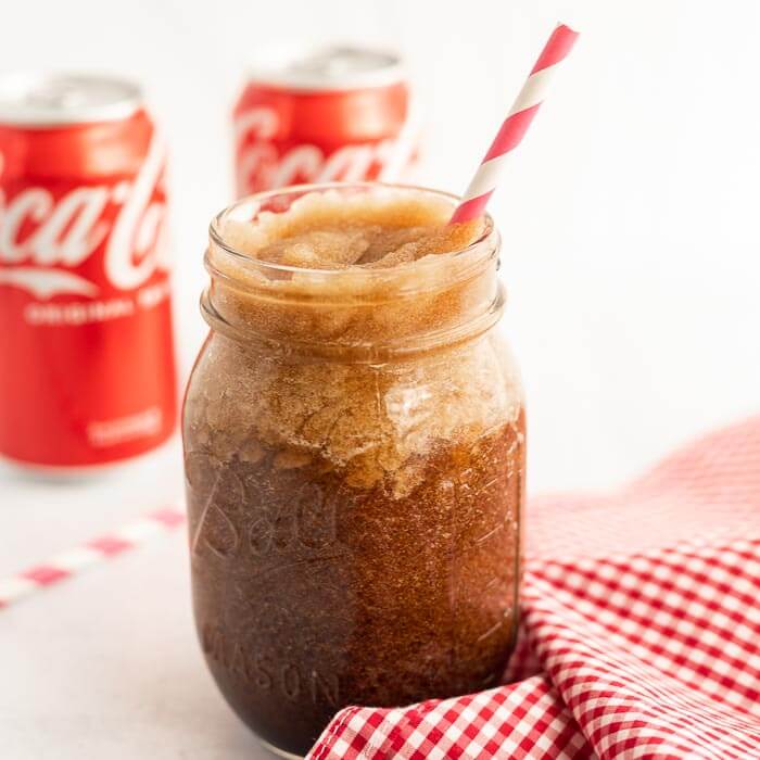 Close up image of a coke slushie in a mason jar with a straw and two cans of coca-cola in the background