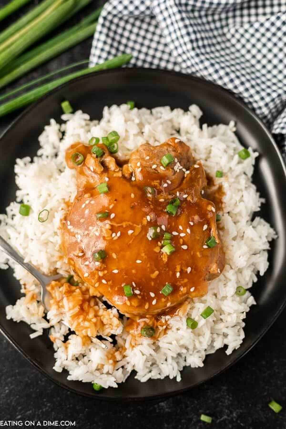 A black bowl filled with white rice, topped with Crock Pot Teriyaki Pork Chops glazed to perfection, garnished with sesame seeds and chopped green onions, with a fork on the side and a checkered napkin in the background.