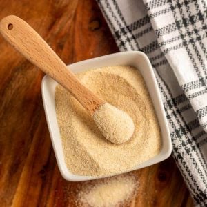 A square white bowl filled with granulated powder, perfect for onion powder substitutes, holds a wooden measuring spoon; a black-and-white checkered towel lies nearby on a wooden surface.