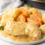 A plate of creamy ranch chicken breast, baby carrots, and potatoes is covered in a thick, golden gravy and garnished with chopped herbs. This comforting instant pot recipe looks tender and inviting.