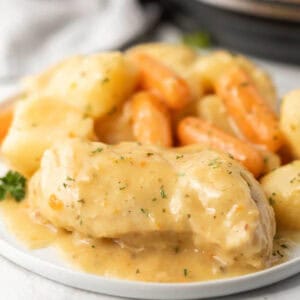 A plate of creamy ranch chicken breast, baby carrots, and potatoes is covered in a thick, golden gravy and garnished with chopped herbs. This comforting instant pot recipe looks tender and inviting.
