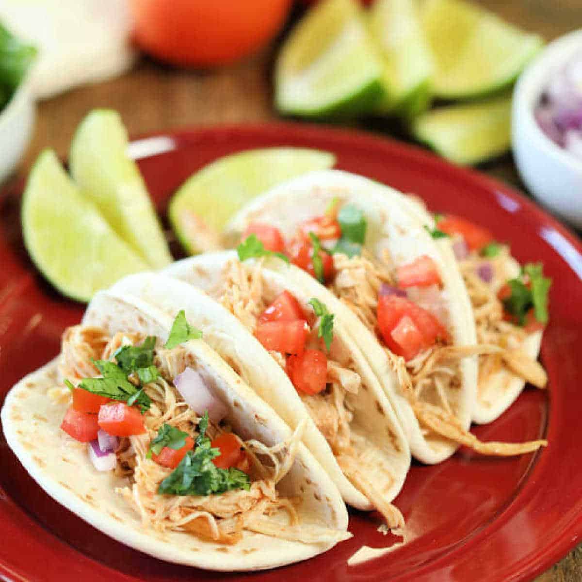 Three tacos filled with shredded chicken, diced tomatoes, and cilantro, served on a red plate. Lime wedges and additional toppings, including onions and green herbs, are arranged around the plate. Instant pot Chicken ranch tacos recipe.