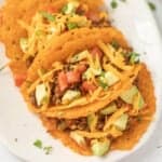 Three tacos with crispy cheese shells, filled with seasoned ground beef, diced tomatoes, avocado, chopped cilantro, and shredded cheddar cheese—this keto street tacos recipe is served on a white plate with scattered herbs.