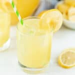 A glass of refreshing Pineapple Lemonade with ice cubes, a green striped straw, and a lemon slice garnish. Extra lemon slices and a blurred drink appear in the background.