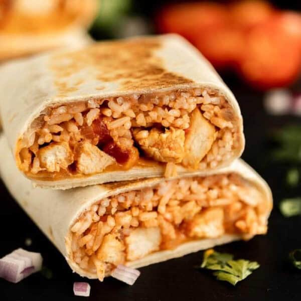 A close-up of a burrito cut in half, showing the delicious filling from a chicken burrito recipe—rice, chicken, beans, and sauce wrapped in a flour tortilla. The burrito is stacked on a dark surface with scattered onions and cilantro.