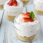 Small glass jars filled with no bake strawberry cheesecake in a jar: a crumbly crust, pink creamy filling, and topped with fresh strawberry slices and a sprig of mint, placed on a light wooden surface.