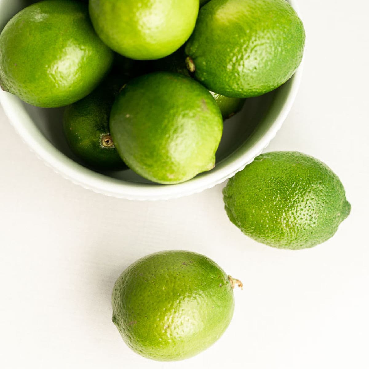 A white bowl filled with fresh green limes sits on a white surface, with three limes placed outside the bowl in the foregroundโperfect for juicing or finding the best substitute for lime juice in your favorite recipes.