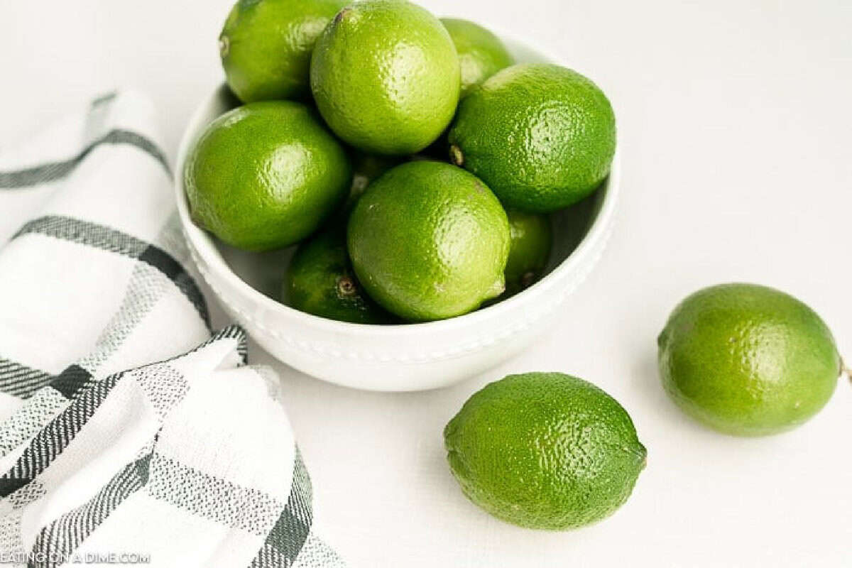 A white bowl filled with fresh green limes sits on a white surface next to a black-and-white checkered kitchen towel, perfect for anyone seeking the best substitute for lime juice, with two limes placed outside the bowl.