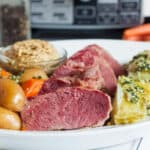 A plate featuring sliced corned beef alongside boiled potatoes, carrots, and cabbage, with a small bowl of mustard. The background shows a kitchen appliance, suggesting preparation for a Crock Pot Corned Beef and Cabbage Recipe.